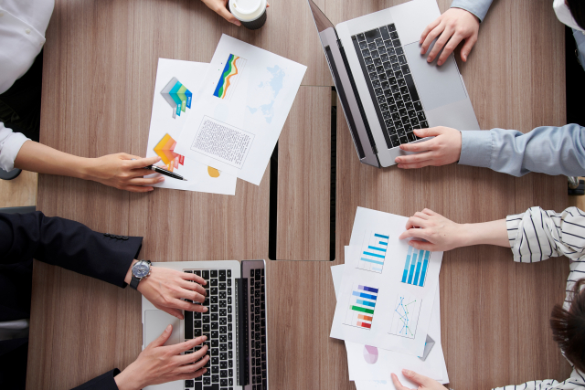 4人の会社員が会議テーブルでPCや資料を見せ、会議している手元の写真