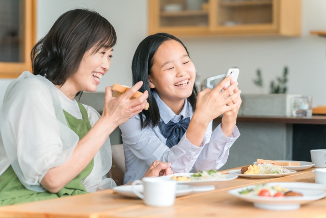 学生服を着た女の子とお母さんが、ご飯を食べながら、スマートフォンを一緒に見ている写真
